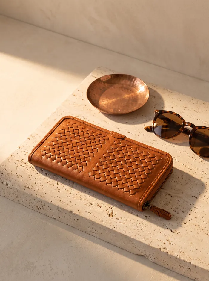 A tan hand-woven leather wallet on travertine stone beside a copper dish