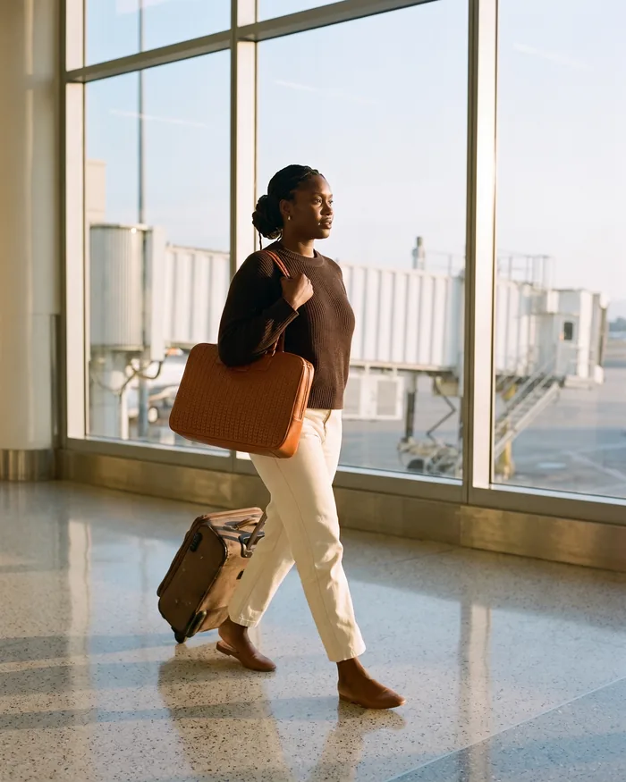 A woman in a deep espresso knit walks an airport departure hall with a tan woven leather tote and a rolling suitcase