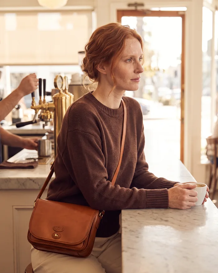A red-haired woman at a marble coffee counter wearing a tan leather saddle crossbody, a cup in her hand