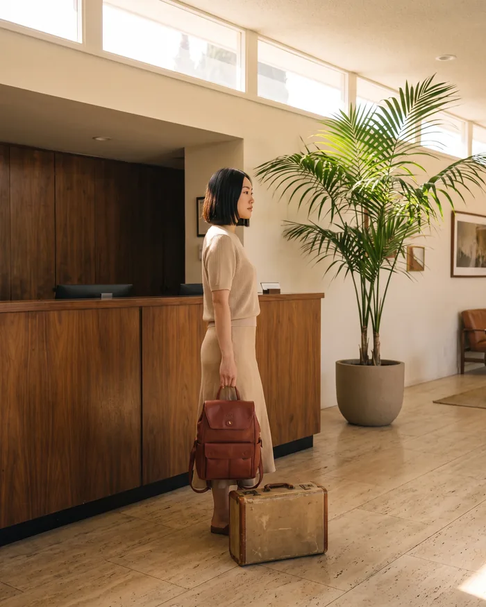 A woman at a hotel reception desk holding a brick leather backpack, a small suitcase at her feet