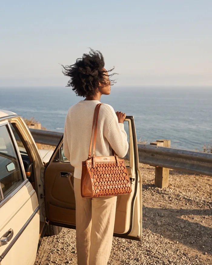 A woman at the open door of a car on a Pacific Coast Highway overlook, a tan woven shoulder bag on her arm